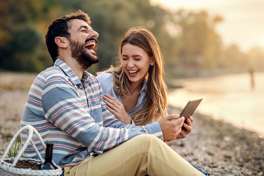 Blog - Young Couple Sitting By The Lake Holding A Tablet And Laughing